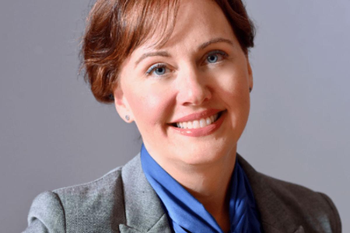 Photo of Kristina Leyden. She has short brown hair and is wearing a grey blazer over a blue shirt with a bow tie.
