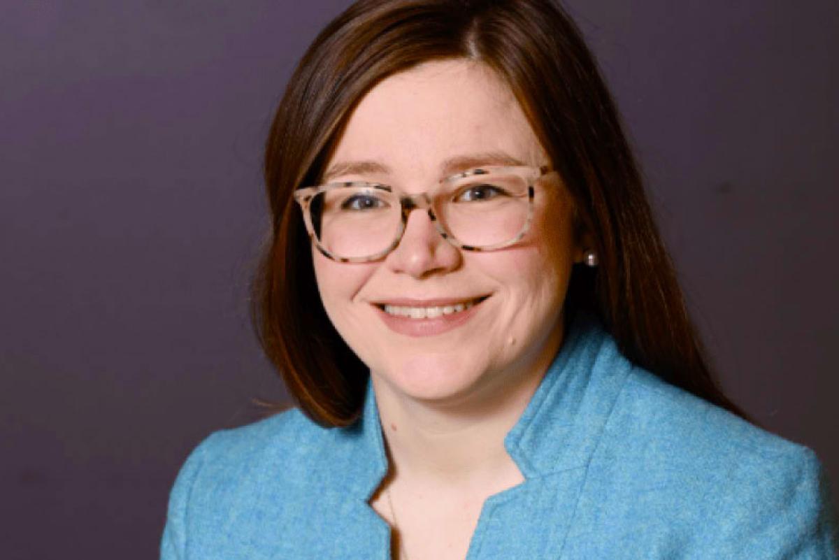 Photo of Lauren Arrigoni. She has long brown hair and glasses. She is wearing a blue blazer over a black top.