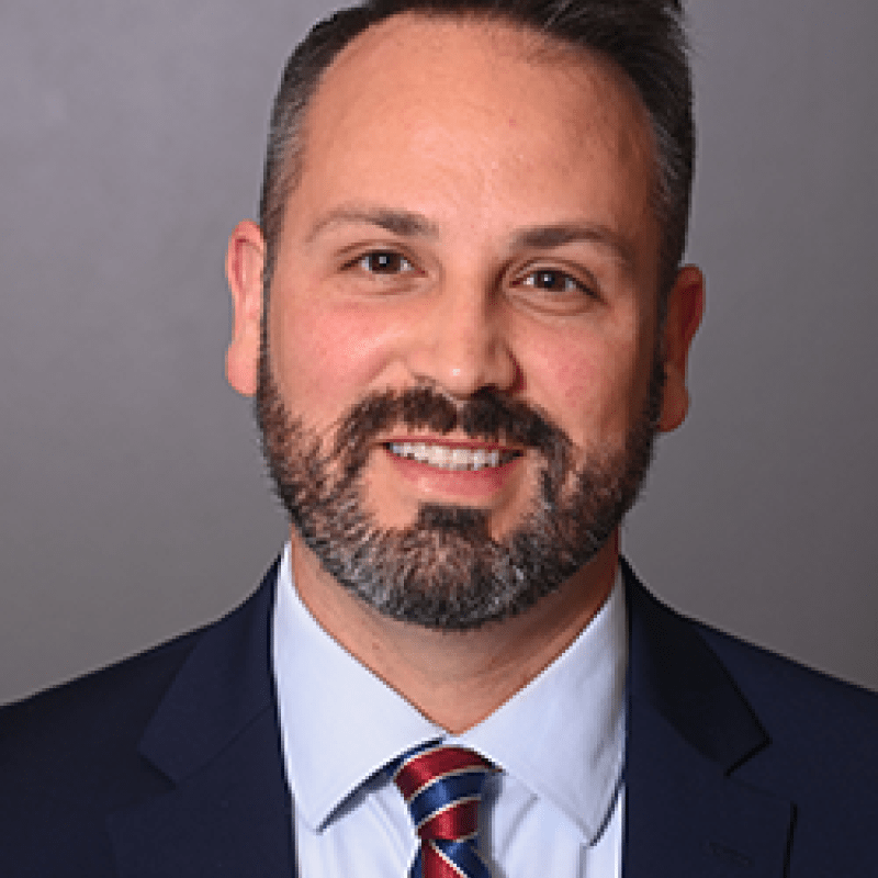 Photo of David Want. He is wearing a navy suit over a light blue shirt and a red and blue tie. He has short brown hair and a beard.