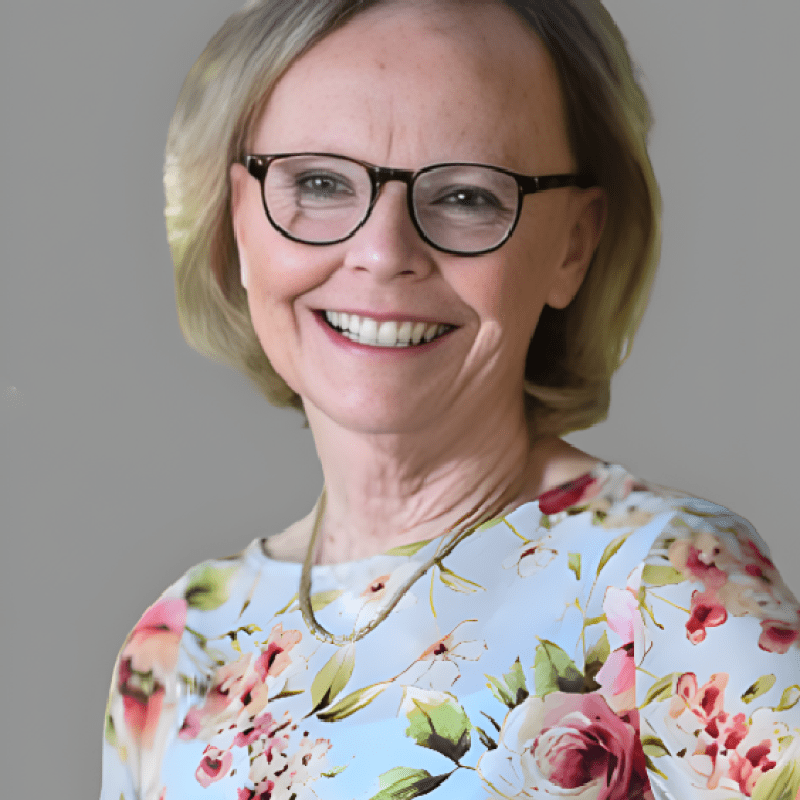 Photo of Elizabeth Hawkins-Walsh. She has bobbed blonde hair and is wearing a blue blouse with large floral prints.