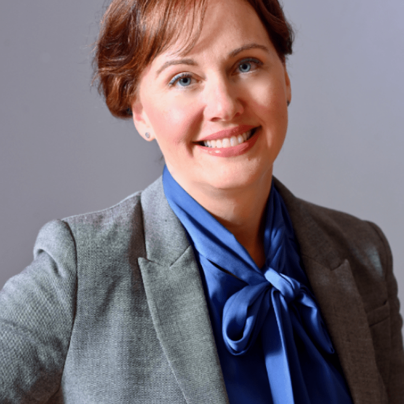 Photo of Kristina Leyden. She has short brown hair and is wearing a grey blazer over a blue shirt with a bow tie.