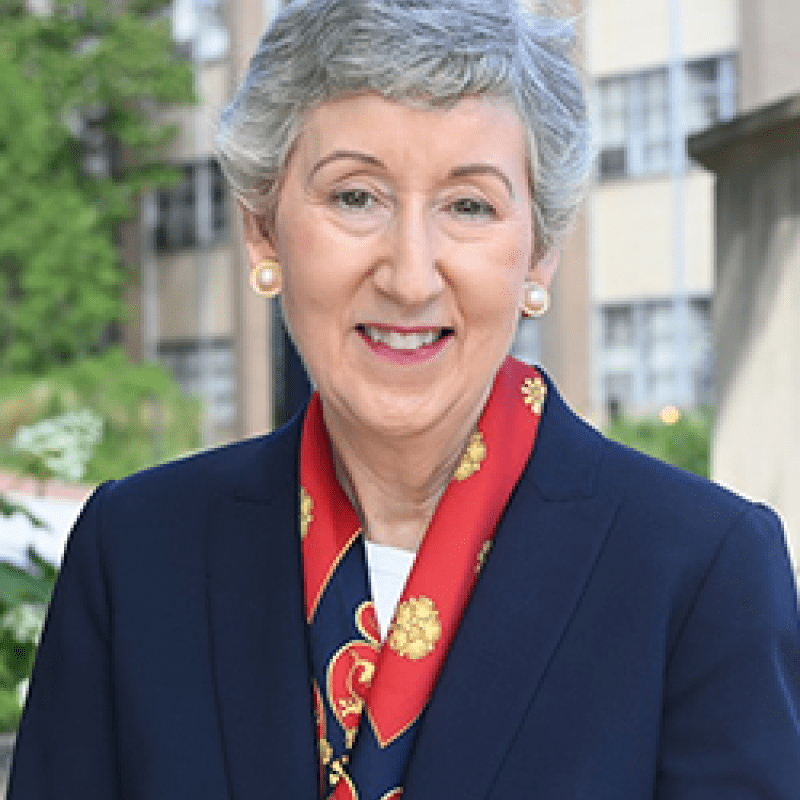 Photo of Marie Nolan outside on Catholic University campus. She has short grey hair. She is wearing a navy blazer over a white shirt and a red silk scarf.