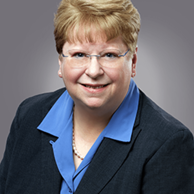 Photo of Janice B. Griffin Agazio. She has blonde hair and glasses and is wearing pearls and a grey blazer with a blue shirt.
