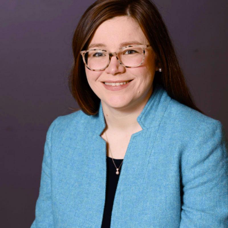 Photo of Lauren Arrigoni. She has long brown hair and glasses. She is wearing a blue blazer over a black top.