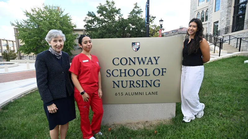 Dean Marie Nolan standing with two Conway School of Nursing students in front of the new sign and building. One student is wearing red scrubs and the other is wearing white pants and a black top. Dean Nolan is wearing a grey and black suit with a skirt.