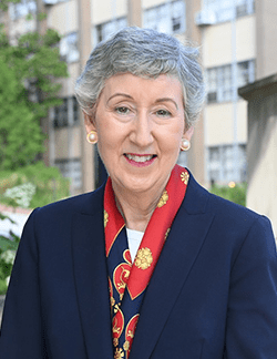 Photo of Marie Nolan outside on Catholic University campus. She has short grey hair. She is wearing a navy blazer over a white shirt and a red silk scarf.